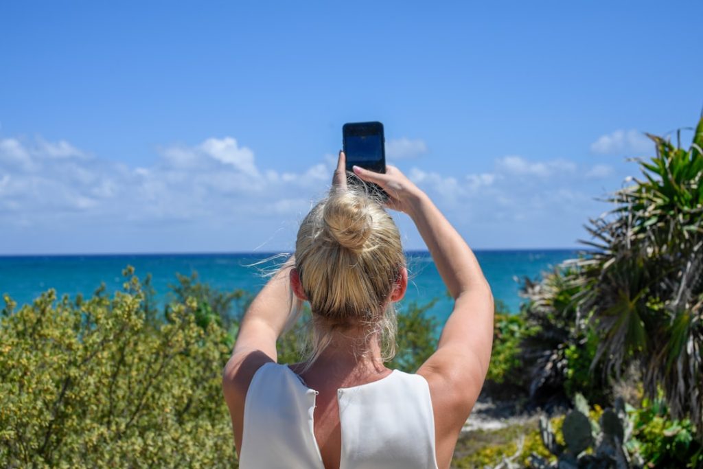 A blonde woman with a hair bun taking a photo of the turquoise ocean with her smartphone.